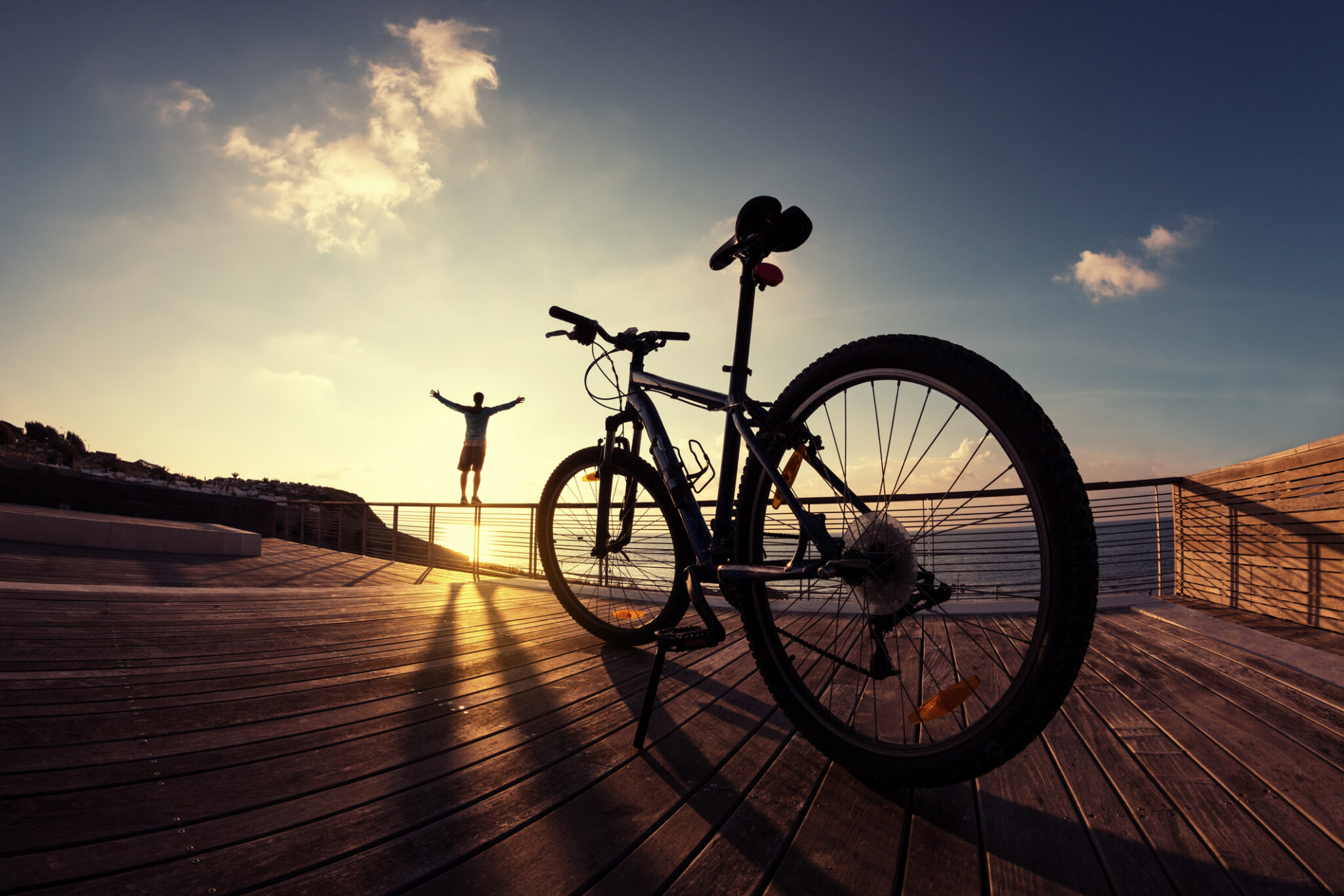 silhouette of young and active sportsman and his mountain bike standing on the railing with outstretched arms near the ocean and looking far away at the sunset